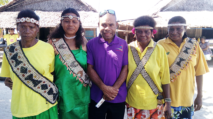 Slade, wearing a green dress of her own creation, is photographed with members of the Weaver's Association that Slade established. They are displaying accessories they threaded from seashells for visitors from the Anglicare Church Partnership Program. Anglicare’s Church Partnership Coordinator, Thompson Yawe, stands with them ©Mavis Tito, 2018