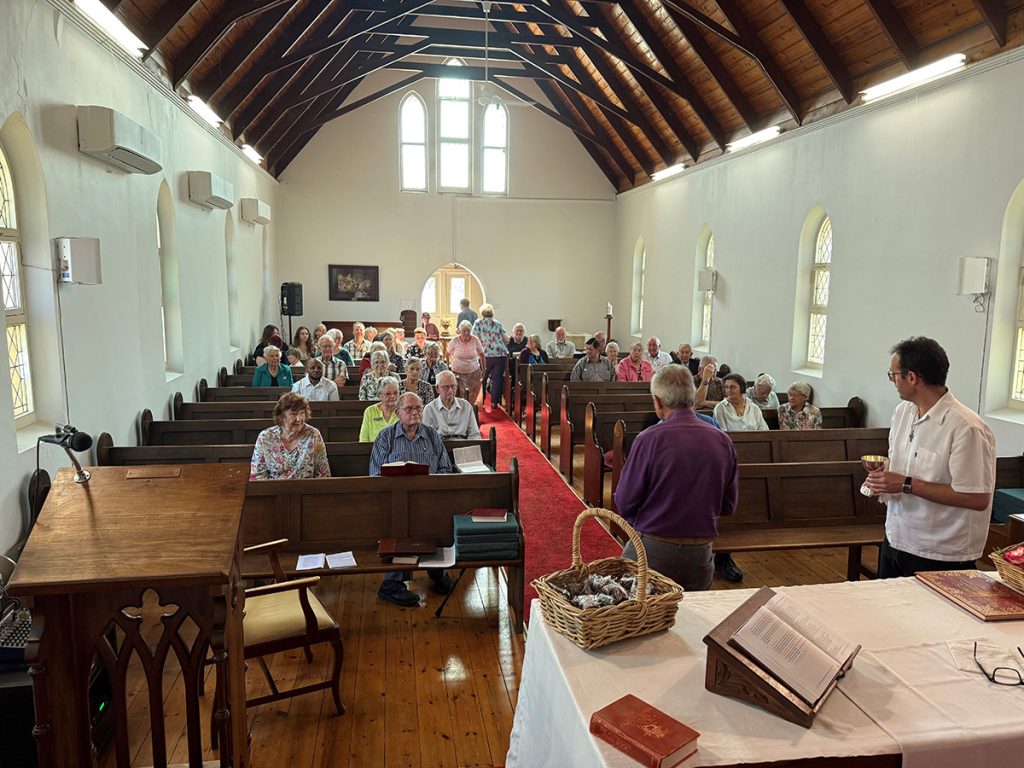 The Rev’d Bruce Hicks (centre purple shirt) invites people forward for communion with the Rev’d Daniel Irvine (far right) assisting © Matt McCue used with permission.